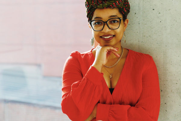 Headshot of Keri. She is wearing an orange top, gold necklaces, white pants, and a green and orange headwrap.