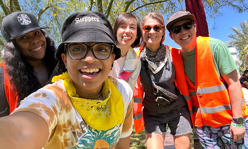 Keri Gary is standing in the front, holding a phone and taking a picture with members of the HR and DEI team at Goldenvoice and parent company AEG. Keri is wearing a black bucket hat, black-rimmed glasses, a yellow scarf, and an orange tie-dye shirt. On the left side of the picture, there is a dark-skinned person wearing a black cap and long black wavy hair. On the right side, there are three individuals wearing bright orange worker vests, all with medium and light skin tones.
