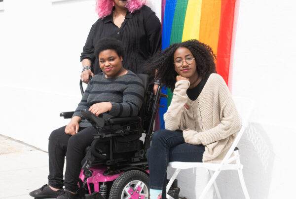 Three Black and disabled folx (a non-binary person holding a cane, a non-binary person sitting in a power wheelchair, and a femme sitting in a chair) casually smile at the camera while a rainbow pride flag drapes on the wall behind them.