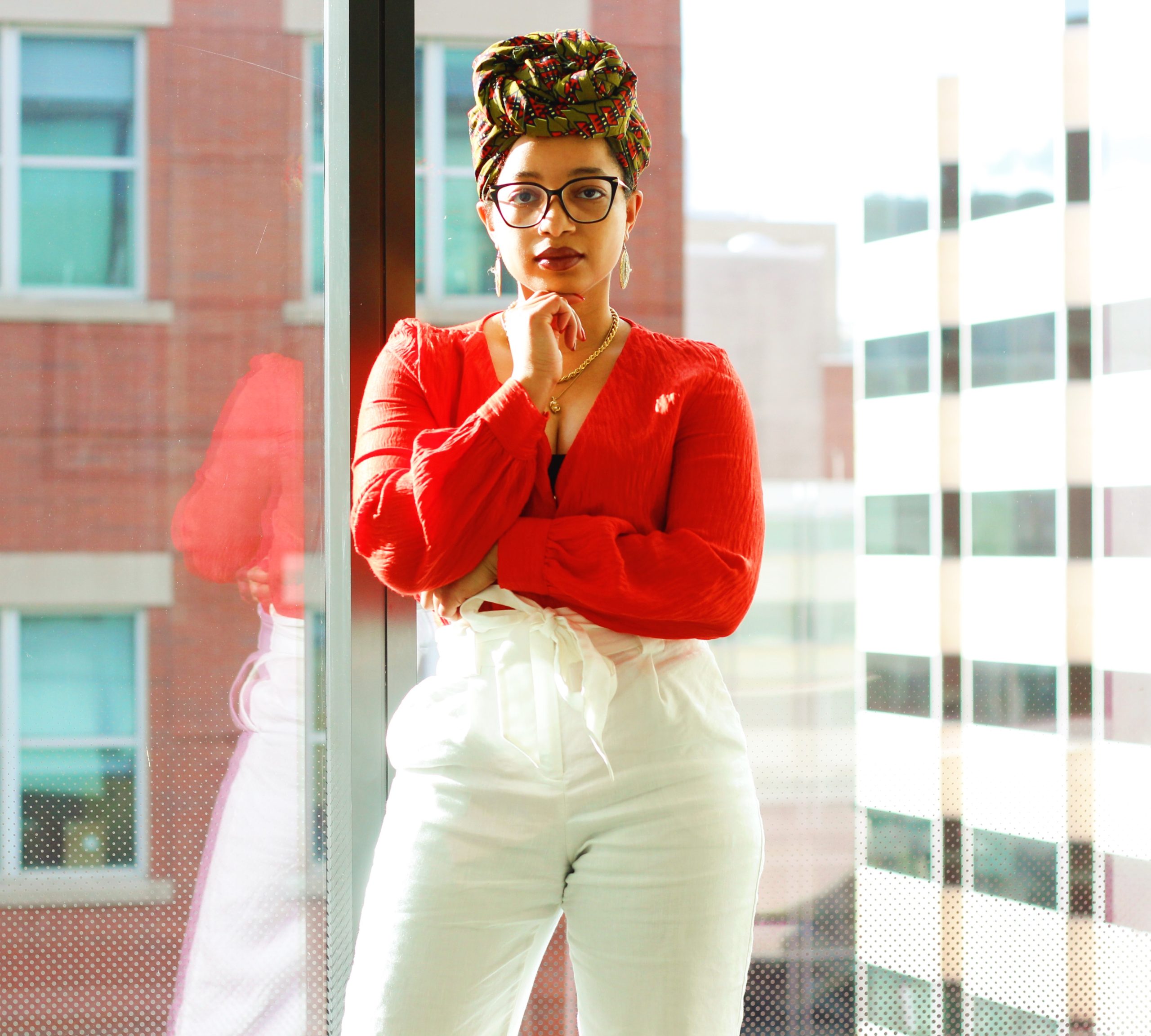 Photo of Keri wearing an orange shirt, white pants, and green and orange headwrap. She appears to be in an office with a large glass window with a couple of building in the background.